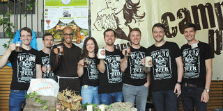 Acht Personen stehen in einheitlichen, schwarzen T-Shirt nebeneinander und haben vor sich auf einem Tisch Hopfen und Malz liegen
