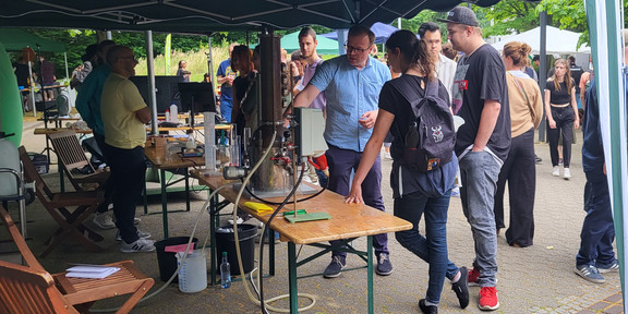 AM Stand der BCI stehen mehrere Besucher und schauen sich die ausgestellten Exponate an. Ein Mitarbeiter erklärt die Funktionsweise