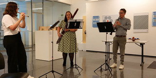 The picture shows three musicians performing during the opening event of "Engineering meets Art" at the Dortmunder U. The first musician is on the left, playing a flute. She is wearing a white T-shirt and black pants. In the middle is another musician playing a bassoon and wearing a green, patterned dress. The third musician is on the right and plays a small trumpet or clarinet. He is dressed in a checked shirt and light-colored pants. In the background you can see some works of art on the walls and a table with various objects on it. The atmosphere is creative and inspiring, in keeping with the theme of the event.