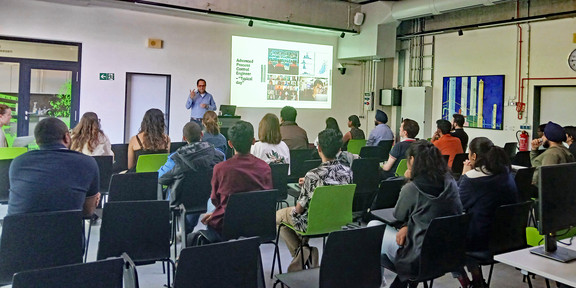 In the BCI Gallery during the event "How to get a job in Germany" Dr. Reinaldo Hernandez presents in front of an audience. On a screen behind him are various pictures and information on the topic of starting a career in Germany. The audience sits in rows on chairs and listens attentively, while some take notes or look at the speaker. In the background, colorful artwork on the walls and technical equipment are visible.