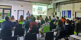 In the BCI Gallery during the event "How to get a job in Germany" Dr. Reinaldo Hernandez presents in front of an audience. On a screen behind him are various pictures and information on the topic of starting a career in Germany. The audience sits in rows on chairs and listens attentively, while some take notes or look at the speaker. In the background, colorful artwork on the walls and technical equipment are visible.