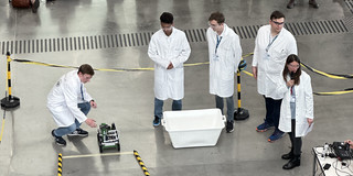 The BCI ChemCar stands on the ground at the starting line. A student kneels next to the vehicle. 4 people in white lab coats are watching him.