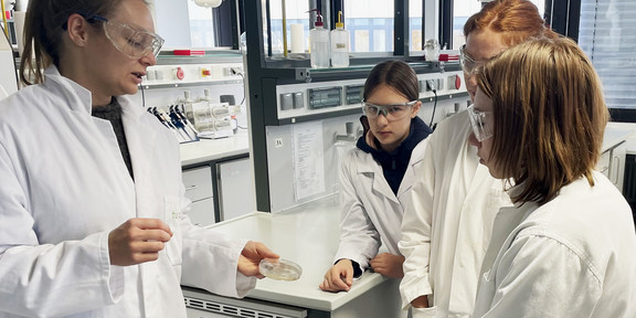  Several participants in white lab coats stand together in a laboratory workshop on the subject of microorganisms. A BCI scientist holds an agar plate in her hand and explains something about the microbes growing on it. The participants listen attentively and look at the plate. Various laboratory equipment and work surfaces used for experiments are visible in the background.