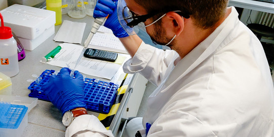 Ein männlicher Wissenschaftler in einem Labor trägt eine Schutzbrille, eine Gesichtsmaske und blaue Einweghandschuhe. Er verwendet eine Pipette, um Proben in einem blauen Reagenzglashalter zu bearbeiten. Auf dem Tisch vor ihm befinden sich Laborutensilien, eine Sprühflasche, ein Taschenrechner und verschiedene Chemikalien. Der Arbeitsplatz ist gut organisiert und modern ausgestattet.