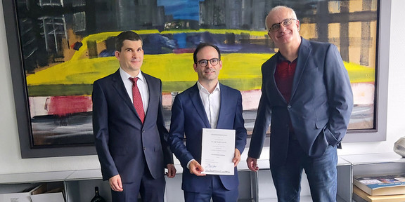 Prof. Lucia präsentiert seine Urkunde, rechts von ihm steht der Prodekan Lehre & Studium, Prof. Nett und links von ihm steht der Rektor, Prof. Bayer, 