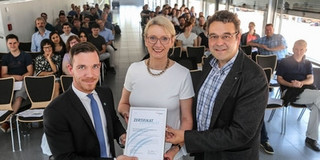 Eine Frau und zwei Männer stehen vor Publikum in einem Raum. Die Frau hält eine Auszeichnung in der Hand