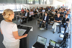 Personen sitzen in einem Raum und hören einer Sprecherin am Rednerpult zu