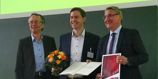 Dennis Vogelsang erhält Carl-Roth-Förderpreis Drei Personen stehen auf einer Bühne nebeneinander. In der Mitte steht ein Mann mit einer Urkunde und einem Blumenstrauss