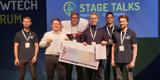The ChemCar competition team celebrates its victory. The members stand proudly on a stage and hold various certificates in their hands. One of the team members is also holding a large cheque showing the prize money. A banner with the title 'Stage Talks' and further information about the competition can be seen in the background.