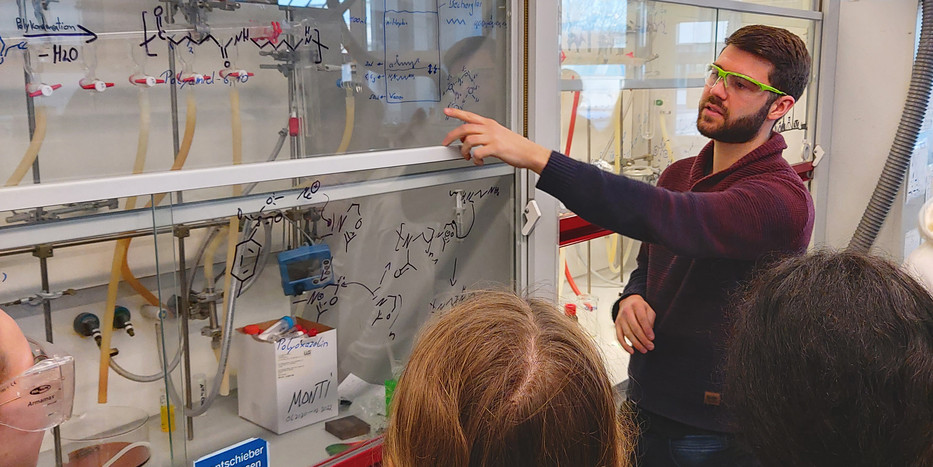 Ein Mitarbeiter erklärt einer Schülergruppe im Labor die Strukturformel von Polymeren