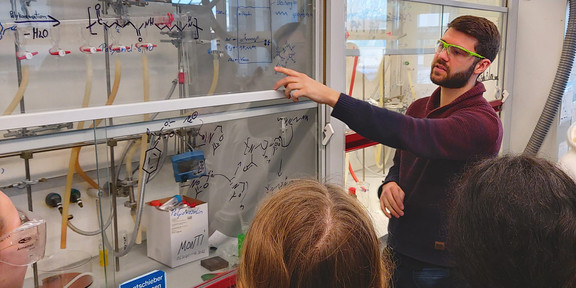 Ein Mitarbeiter erklärt einer Schülergruppe im Labor die Strukturformel von Polymeren