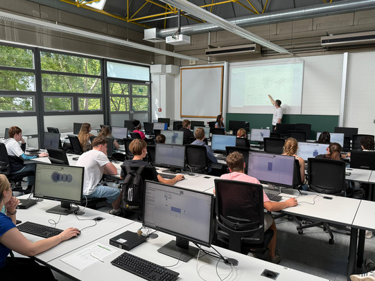 The picture shows a classroom with several people working on computers. A presentation is displayed on the wall while one person stands at the front and explains. The audience is sitting in ergonomic chairs and looking at their screens or at the presentation.
