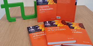 Three books with orange cover are lying on a pile, two books are standing behind it, to the left of it the green TU Dortmund logo