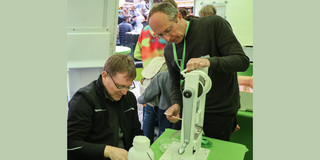 Dr. Kerzel, the Managing Director of BCI, explains to a visitor how a tablet press works in a tent at TU Dortmund University during the DortBunt event. The visitor observes attentively while Dr. Kerzel performs a demonstration with the device. Other people and stands at the event can be seen in the background.