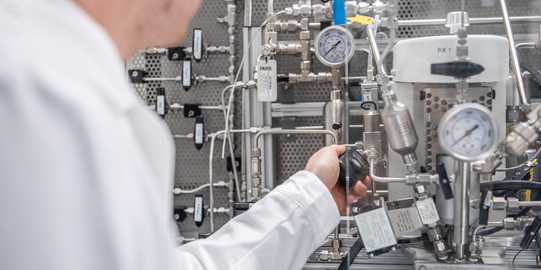 A chemical engineer in a white lab coat operates a complex reactor system in a technical laboratory. The focus is on a variety of pipes, valves, measuring devices and pressure gauges that are part of a larger experimental setup. The engineer holds a valve handle and checks the settings on the machines. The environment looks industrial and highly technical.