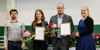 Zwei Männer und zwei Frauen stehen vor einer Tafel nebeneinander. Die Personen in der Mitte halten Urkunden und Blumensträusse in den Händen