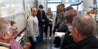 Mann mit Schutzbrille und Handschuhen empfängt  Besucher im Labor