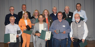 The picture shows a group of people who have received the silver diploma certificate of honor from the Department of Biological and Chemical Engineering at Dortmund University of Technology. A total of eleven people are pictured standing in two rows. Some of them are holding the award in their hands and smiling at the camera. The people are predominantly middle-aged to elderly. There are six people in the front row and five in the back. The awards consist of a certificate and a small object. The event takes place in an interior room in front of a gray curtain.