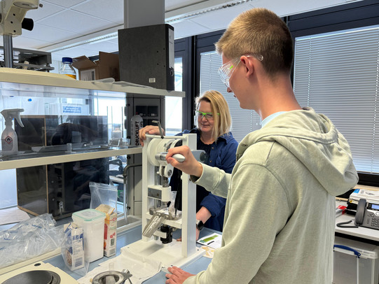 he picture shows a person working on a crank-operated tablet press in a laboratory. The person is holding the crank in their hand and concentrating on the device. Various laboratory materials and utensils used for experiments are visible in the background. The room is brightly lit and conveys a working atmosphere.