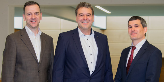 The picture shows three men in formal dress smiling and looking at the camera. Pictured from left to right are: Prof. Hannsjörg Freund in a brown suit, Prof. Norbert Kockmann in a dark blue suit and Prof. Markus Nett in a dark blue suit with a red tie. They are standing next to each other in a modern building.