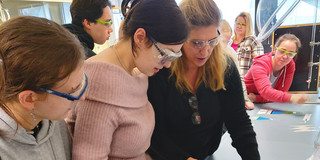 Eine Gruppe von Menschen trägt Schutzbrillen und arbeitet an einem Experiment. Eine Mitarbeiterin der Fakultät BCI erklärt Schülern, wie Pfefferminzöl aus der Pfefferminzpflanze extrahiert wird. Im Hintergrund sind weitere Personen zu sehen, die sich auf die Arbeit konzentrieren.