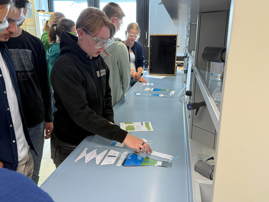 The picture shows a group of people standing at a table in a laboratory. One person is actively sorting various materials and arranging them on the table. In front of her are several colorful pieces of paper containing colorful graphics and information. In the background, other participants can be seen observing attentively or chatting. The room is well lit, with large windows through which daylight streams in. 