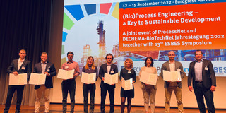 9 person are standing on a stage, 8 of them are holding a certificate. All are elegantly dressed. In the background you can see a projection with the text "Welcome to Eurogress Aachen. September 12-15, 2022 BioProcess Engineering - a Key to Sustainable Development. A joint event of ProcessNet and DECHEMA-BioTechNet Annual Conference 2022 together with 13th ESBES Symposium.