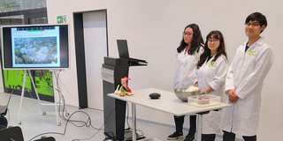 KimchiZubereitung vor dem Webcam in der BCI galerie, die 3 Studierenden stehen hinter einem Tisch. Auf dem Tisch liegen die benötigten Zutaten. 