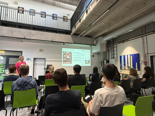The picture shows a group of people in a seminar room looking at a presentation on the wall. One person stands at the front and presents, while the audience sits on chairs and follows attentively. The chairs are green and black, and some pictures on the wall and technical equipment are visible in the background. The room has high ceilings and large windows that create a bright atmosphere.