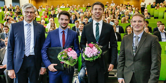Vier Männer stehen nebeneinander im Hörsaal, im Hintergrund sitzt Publikum. Die beiden Männer in der Mitte halten Blumensträusse in den Händen