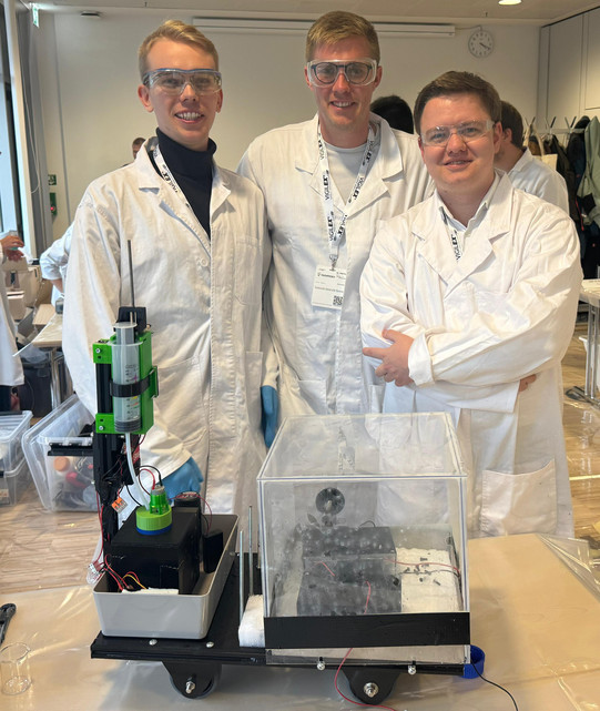 The ChemCar competition team proudly presents their self-developed vehicle. The team members wear white lab coats and stand around the vehicle, which is placed on a table. The vehicle consists of various components, including a transparent box that makes the technical elements visible, as well as an engine and other mechanical parts. Other participants in the competition can be seen in the background.