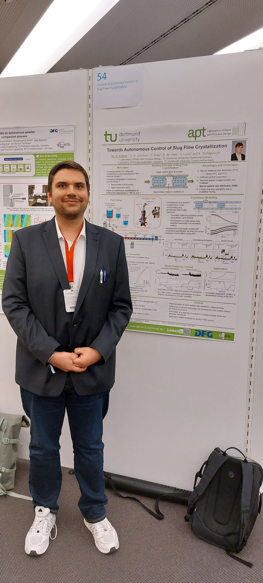 Maximilian Kattner steht vor seinem Gewinnerposter mit dem Titel 'Towards Autonomous Control of Slug Flow Crystallization'. Er trägt einen dunklen Anzug und eine rote ID-Karte um den Hals. Das Poster zeigt verschiedene Diagramme, Grafiken und Textabschnitte, die sich mit der autonomen Kontrolle von Kristallisationsprozessen beschäftigen. Im Hintergrund sind weitere Posterständer zu sehen.