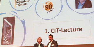 Prof. Norbert Kockmann hält 1. CIT-Lecture Ein Mann und eine Frau stehen auf einer Bühne, im Hintergrund eine Beamerwand mit Präsentation