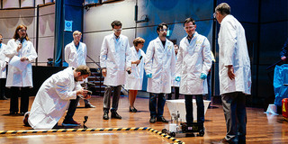 ChemCar-Team 2018 Acht Personen in weissen Kitteln stehen auf einer Bühne und führen das Modell vor
