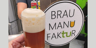 Ein Glas mit bernsteinfarbenem Bier, bedeckt von einer dichten Schaumkrone, wird vor einem Ausschank präsentiert. Im Hintergrund ist ein Schild mit der Aufschrift 'BRAU MANU FAKtur' zu sehen, wobei der Buchstabe 'U' als stilisiertes Bierglas dargestellt ist. Das Bier wird bei der Verkostung der Brau-AG präsentiert, die jährlich ein neues Bier an der Fakultät entwickelt.