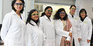 Sechs frauen stehen in weissen Kitteln und Schutzbrillen nebeneinander in einem Labor