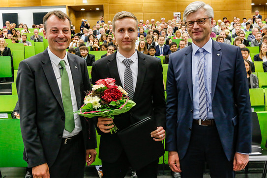 Drei Männer stehen im Hörsaal vor Publikum, der Mann in der Mitte hat einen Blumenstrauss in der Hand