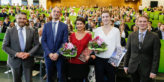 Fünf Personen stehen im Hörsaal nebeneinander, im Hintergrund Publikum.