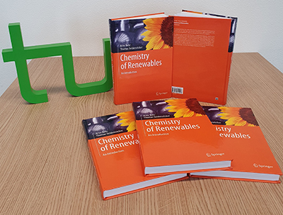 Three books with orange cover are lying on a pile, two books are standing behind it, to the left of it the green TU Dortmund logo