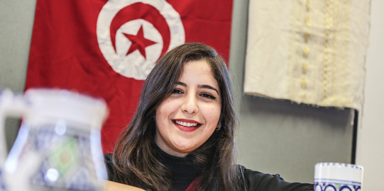 Eine internationale Studierende lächelt in die Kamera. Im Hintergrund hängt eine Tunesienflagge.
