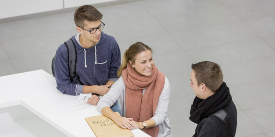 Three people are standing in front of a service desk. The person in the middle is holding an envelope.