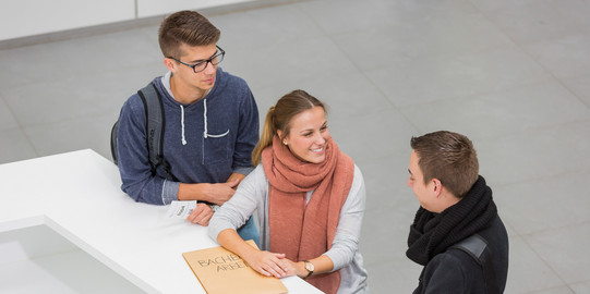 Three people are standing in front of a service desk. The person in the middle is holding an envelope.