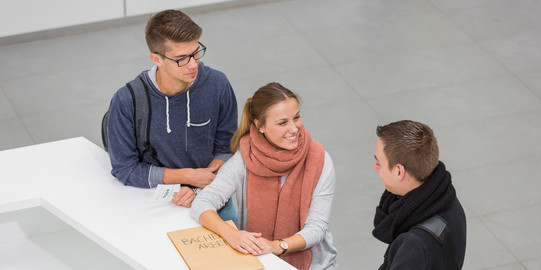 Three people are standing in front of a service desk. The person in the middle is holding an envelope.