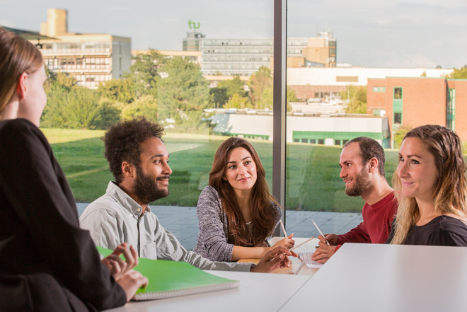 Fünf Studierende sitzen an einem Tisch mit Stiften in der Hand und Papier vor sich und sehen sich an. Im Hintegrund sieht man durch eine große Glasscheibe eine Wiese, Bäume und andere Gebäude. Auf einem Gebäude ist das grüne TU-Logo zu sehen.