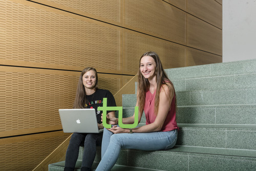 Two students are sitting on a stair. One is holding a laptop and the other the logo of TU Dortmund University