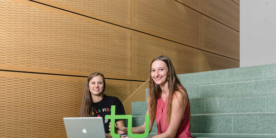 Zwei Studentinnen sitzen mit Laptop und TU-Logo auf einer Treppe.