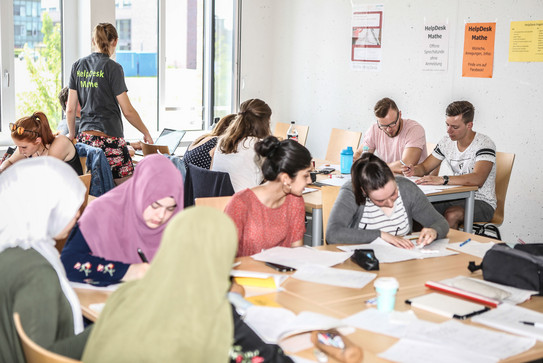 Mehrere Personen sitzen an Tischen und haben Unterlagen vor sich. Eine Person steht und trägt ein T-Shirt mit der Aufschrift "HelpDesk-Mathe".